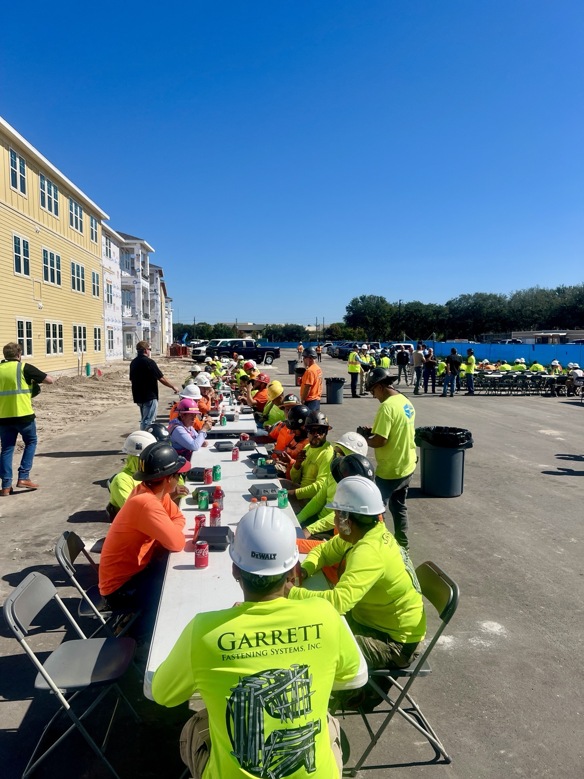 A large outdoor workplace gathering sharing a meal at long tables